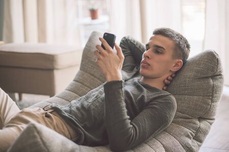 Handsome teenage guy with dental braces using smartphone while relaxing on modern couch in living room interior. Young man resting in soft chair at home.の写真素材