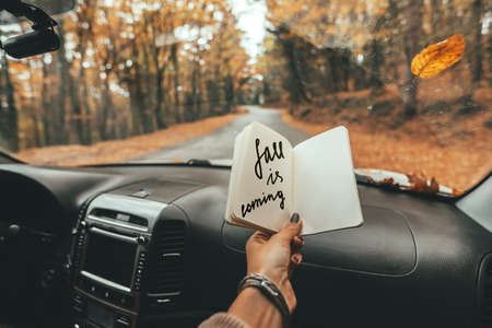 Road trip looking inside of car. Woman hand holding notebook with Fall Is Coming text over windshield. Freedom travel concept. Autumn weekend to the forest.の写真素材