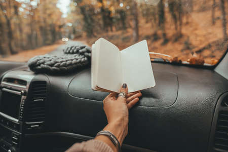 Road trip looking inside of car. Woman hand holding notebook with blank pages over windshield. Freedom travel concept. Autumn weekend to the forest.の写真素材