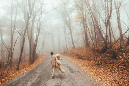 Woman with big plush teddy bear toy walking away on road in magic forest one foggy autumn dayの写真素材