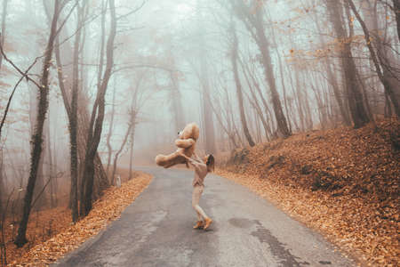 Woman with big plush teddy bear toy walking away on road in magic forest one foggy autumn dayの写真素材