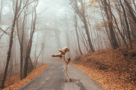 Woman with big plush teddy bear toy walking away on road in magic forest one foggy autumn dayの写真素材