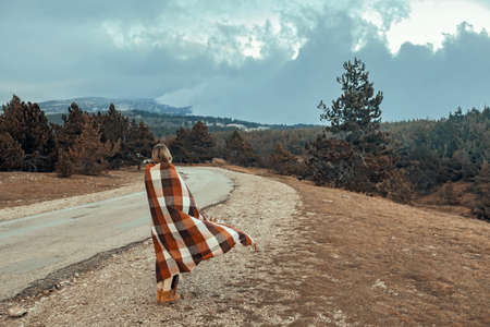 Woman wrapped in warm blanket standing on road and looking away in mountains in cold bad weatherの写真素材