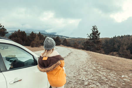 Woman standing by the car and looking away on road in mountains in cold bad weatherの写真素材