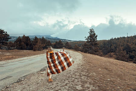 Woman wrapped in warm blanket standing on road and looking away in mountains in cold bad weatherの写真素材