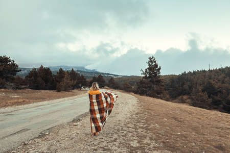Woman wrapped in warm blanket standing on road and looking away in mountains in cold bad weatherの写真素材