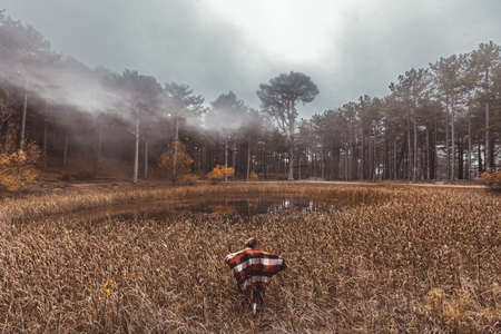 Woman traveller wrapped in plaid blanket relaxing by the lake in autumn in woods in cold fog weatherの写真素材
