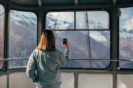 Young traveler taking photo of mountains on her smartphone through sky lift window. Student girl spending her holidays in nordic country.の写真素材