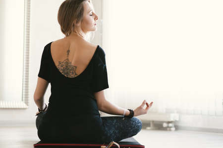 Young woman with mandala tattoo on her back using aroma sticks while doing stretching exercise by mirror on floor mat in bright yoga class room.の写真素材