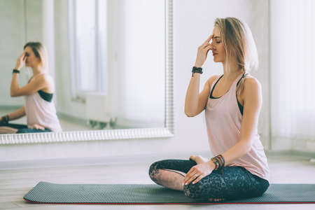 Young woman sitting in yoga pose and meditating by mirror on floor mat in bright class room.の写真素材