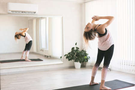 Young woman doing stretching exercise by mirror on floor mat in bright yoga class room.の写真素材