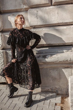 Summer outdoor portrait of young elegant woman wearing black lace maxi dress and autumn leather boots posing in the city street. Natural sun light. City buildings behind the model.の写真素材