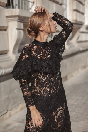 Summer outdoor portrait of young elegant woman wearing black lace maxi dress posing in the city street. Natural sun light. City buildings behind the model.の写真素材