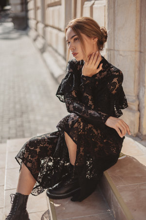 Summer outdoor portrait of young elegant woman wearing black lace maxi dress and autumn leather boots posing in the city street. Natural sun light. City buildings behind the model.の写真素材