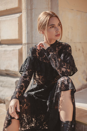 Summer outdoor portrait of young elegant woman wearing black lace maxi dress posing in the city street. Natural sun light. City buildings behind the model.の写真素材