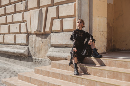 Summer outdoor portrait of young elegant woman wearing black lace maxi dress and autumn leather boots posing in the city street. Natural sun light. City buildings behind the model.の写真素材
