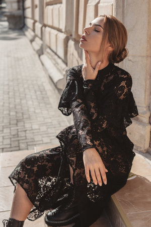 Summer outdoor portrait of young elegant woman wearing black lace maxi dress and autumn leather boots posing in the city street. Natural sun light. City buildings behind the model.の写真素材