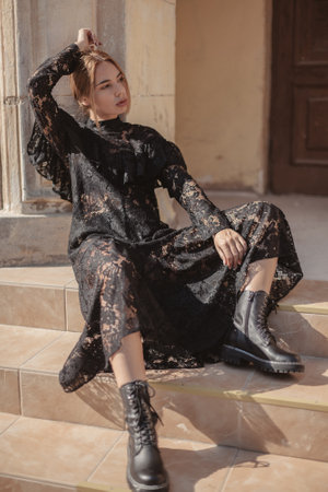 Summer outdoor portrait of young elegant woman wearing black lace maxi dress and autumn leather boots posing in the city street. Natural sun light. City buildings behind the model.の写真素材