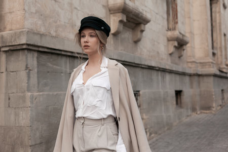 Young stylish beautiful woman wearing white shirt, beige textile coat and black cap posing outdoors against street wall. Trendy casual autumn outfit. Street fashion.の写真素材