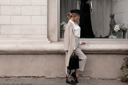 Young stylish beautiful woman wearing white shirt, beige textile coat and pants, black cap and lofers, holding purse posing outdoors against street wall. Trendy casual autumn outfit. Street fashion.の写真素材