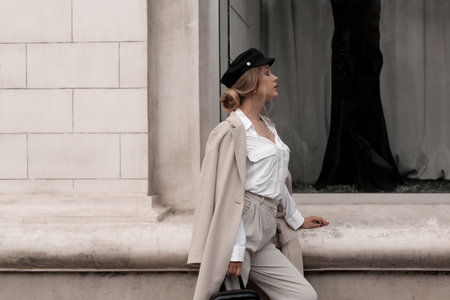 Young stylish beautiful woman with elegant hairstyle wearing white shirt, beige textile coat and black cap posing outdoors against street wall. Trendy casual autumn outfit. Street fashion.の写真素材