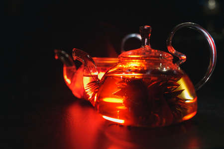 Glass teapot filled with flowers and berries, green tea. Appetizing beautiful drink in orange tones on a black backgroundの写真素材