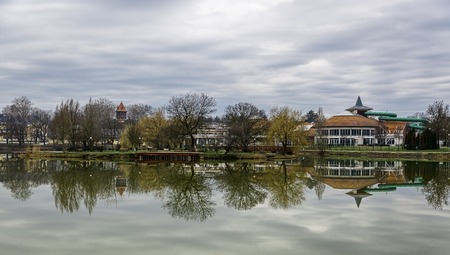 Beautiful lake with mirror reflections in clear water on cloudy day. Tranquil landscape with lake, houses, cloudy sky, and trees reflected symmetrically in the water. Nyiregyhaza, Hungaryの写真素材