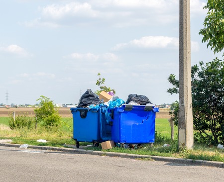 Overflowing blue plastic waste cans with black garbage bags, cardboard boxes and other garbage...