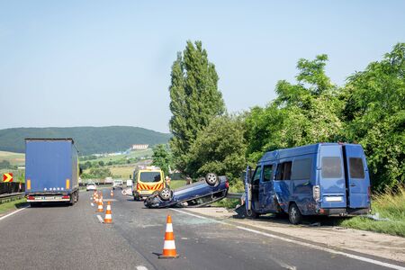 Car crash accident on highway. Damaged blue minibus after collision, overturned car and ambulance car on roadside. Traffic cones at accident siteの写真素材