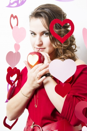 Attractive young girl standing among ornaments from paper hearts and holds a ripe apple with carved heart symbolの写真素材