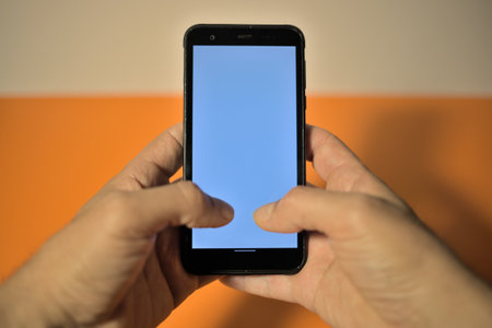 Smartphone held with right hand in foreground display colored purple blank on right background orange and white copy space on leftの写真素材