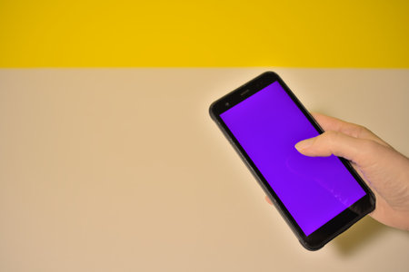 Smartphone held with right hand in foreground display colored purple blank on right background orange and white copy space on leftの写真素材