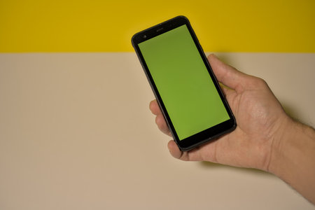 Smartphone held with one hand in the foreground displaying colored green blank on the right against an orange background and white copy space on the leftの写真素材