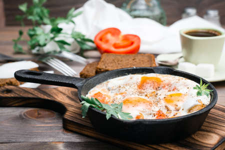 Ready-to-eat breakfast: shakshuka from fried eggs with tomatoes and parsley in a pan, bread with butter and coffeee on a wooden tableの写真素材