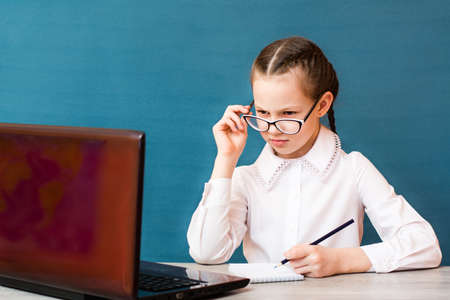 The girl in glasses works at a laptop and makes notes in a notebook at the table. Distance learning...