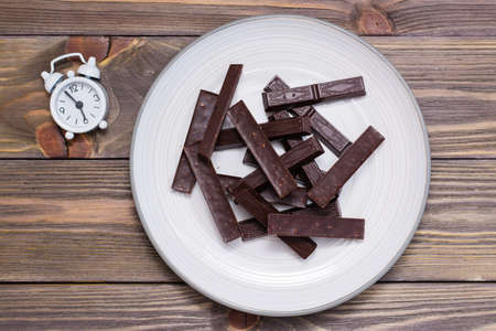 A pile of molded pieces of chocolate on a plate and an alarm clock on a wooden table. The concept of...