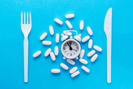 White oval pills, alarm clock, knife and fork on a blue background. Top view. The concept of taking medication at the right time.の写真素材