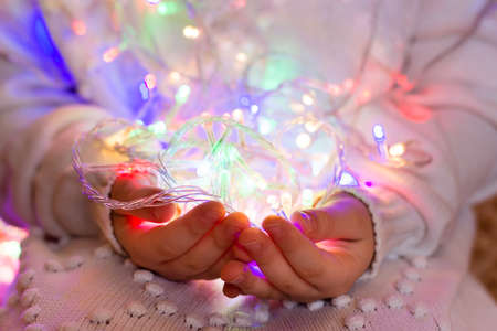 Christmas garland of multi-colored led-bulbs in the hands of a child dressed in a white knitted sweater. The concept of comfort, warmth, the advent of Christmasの写真素材