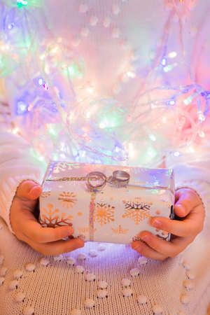 A gift wrapped in a festive packaging in the hands of a child in a white knitted sweater against a...