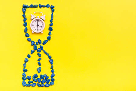 Alarm clock inside an hourglass made of decorative stones on a yellow background. Leaking time...