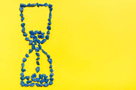 Hourglass from blue decorative stones on a yellow background. Leaking time concept