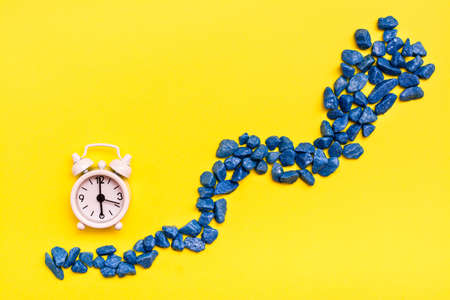 White alarm clock and a road made of decorative stones on a yellow background. Leaking time concept