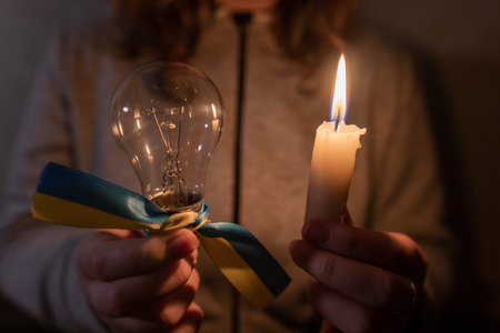 blackout. energy crisis. power outage concept. The girl holds in her hands a burning candle and an electric light bulb with a yellow-blue ribbon (the national symbol of Ukraine)の写真素材