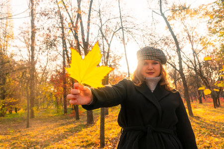 Girl in a black coat holds a yellow leaf in her hands. autumn forest. Sunny day. Autumn walks in the forest. connection with natureの写真素材