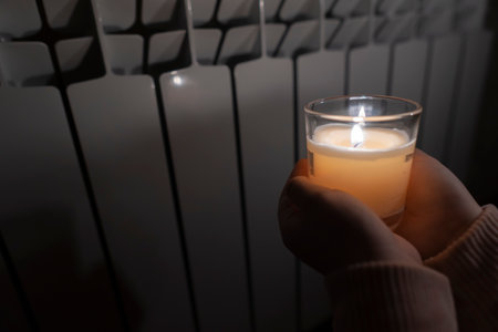 A girl holds a candle in a glass cup in her hands against the background of a household heating battery (close-up). power outage concept. blackout. energy crisisの写真素材