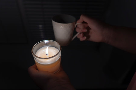 blackout. energy crisis. power outage concept. The girl holds a candle in one hand and a cup of tea in the other hand against the background of a window with blinds (close-up)の写真素材