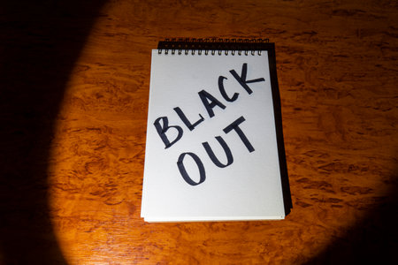 <p>Notepad with the inscription <strong>Blackout</strong> on a wooden table, illuminated by a...