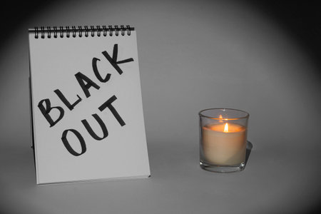 <p>Notepad with the inscription <strong>Blackout</strong> and a burning candle in a glass on a gray...