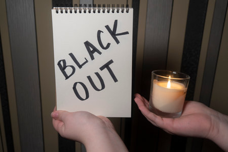A notepad with the inscription "Blackout" in one hand and a burning candle in the other hand (close-up). power outage concept. energy crisis. Destruction of infrastructureの写真素材