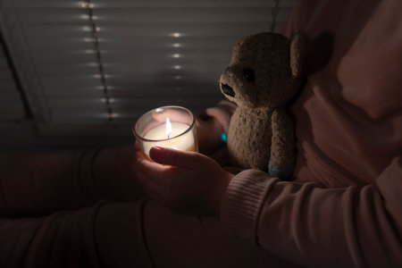 A girl is in a dark room near the window and holds a candle and a teddy bear in her hands (close-up). power outage concept. blackout. energy crisisの写真素材